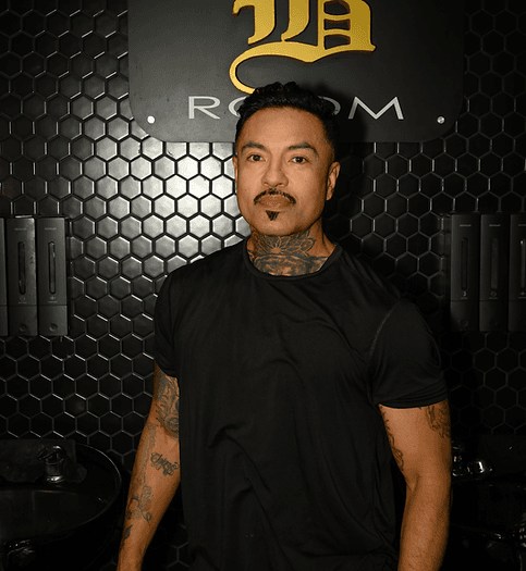 Omar, professional barber with tattoos wearing black shirt, standing in modern barbershop with hexagon wall